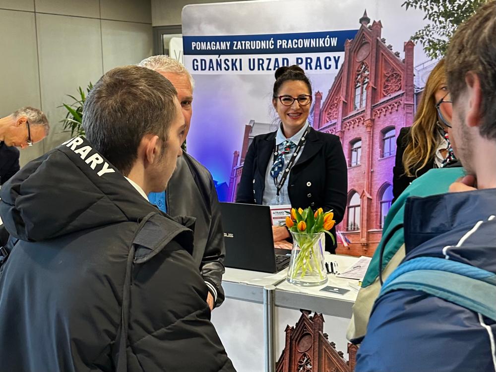 Uśmiechnięta kobieta przy stoisku promocyjnym Gdańskiego Urzędu Pracy. Dookoła niej stoją inne osoby.